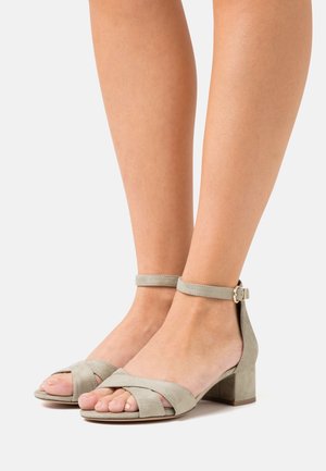 Pieds portant des sandales à talons blocs ouverts en daim gris clair avec des brides à la cheville et des boucles dorées sur fond blanc.