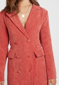 Dvouřadový kordový kabát v jemné korálové barvě, s vertikálně vroubkovanou texturou, s klopami a čtyřmi vzorovanými knoflíky na přední straně.