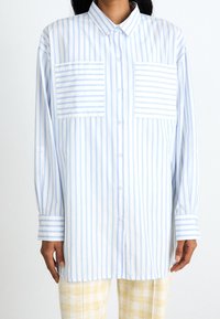 Světle modrá a bílá proužkovaná košile na knoflíky s dvěma předními kapsami, dlouhými rukávy a volným střihem; látka vypadá hladká a prodyšná.