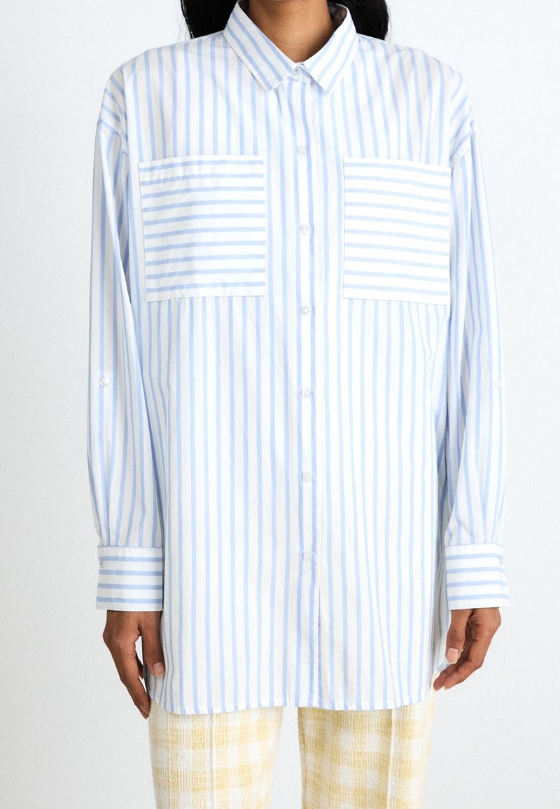 Světle modrá a bílá proužkovaná košile na knoflíky s dvěma předními kapsami, dlouhými rukávy a volným střihem; látka vypadá hladká a prodyšná.