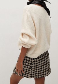 Maglione oversized color crema con texture a coste, abbinato a una mini gonna a scacchi in bianco e nero. Sciarpa nera attorno al collo.