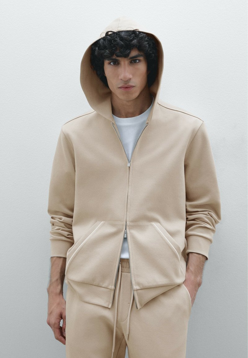 Haut à capuche beige avec fermeture éclair, doté de grandes poches avant et d'un tissu texturé ; superposé sur un t-shirt blanc ; coupe décontractée.