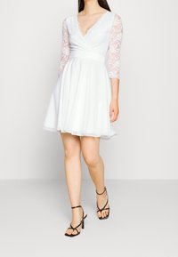 Weißes Kleid mit einem enganliegenden Oberteil, Spitzendrei-Viertel-Ärmeln und einem ausgestellten Rock. Der Stoff ist glatt mit einem dezenten Glanz.