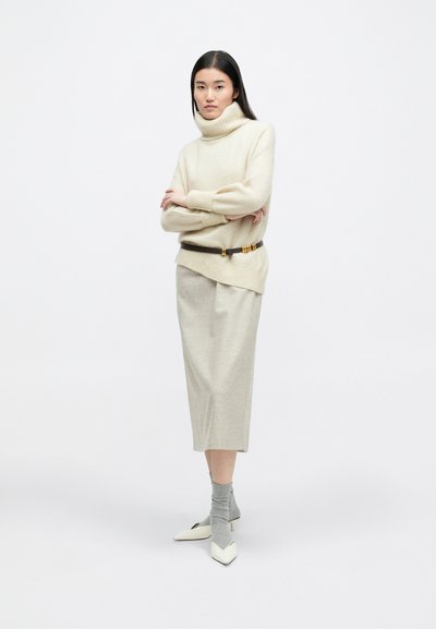 Μπεζ ζιβάγκο πουλόβερ με ριγωτούς καρπούς, συνδυασμένο με μπεζ μίντι φούστα, καφέ ζώνη με χρυσές λεπτομέρειες και λευκά ψηλοτάκουνα παπούτσια με κοφτή μύτη. Γκρι κάλτσες.