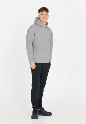 Adolescent debout, portant un sweat à capuche gris, un pantalon de survêtement noir et des baskets noires, sur un fond blanc.