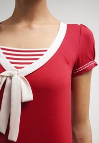 Mujer que lleva una blusa roja con ribetes y un lazo blancos, superpuesta a una camisa de rayas rojas y blancas con mangas cortas y abullonadas.
