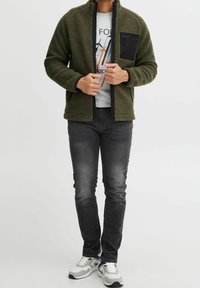 Oliwkowa polarowa kurtka z wysokim kołnierzem, zapinana na zamek, z czarną kieszenią na piersi, w połączeniu z szarymi dżinsami i szarymi trampkami.