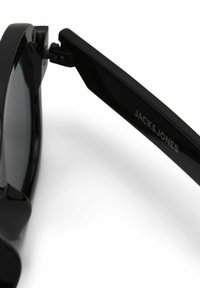 Βραχίονας γυαλιών ηλίου Black Jack & Jones συνδεδεμένος με μαύρο σκελετό με μεντεσέ, εμφανίζεται σε λευκό φόντο.