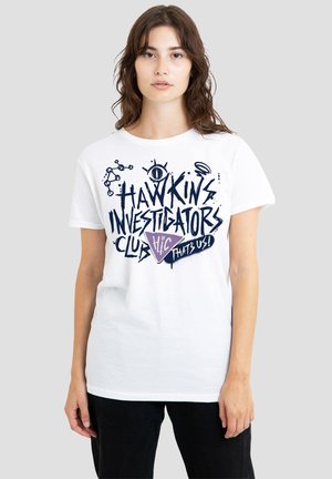 Junge Frau trägt weißes T-Shirt mit dunkelblauem Text und Grafik „Hawkins Investigators Club“ und steht vor einem schlichten hellen Hintergrund.