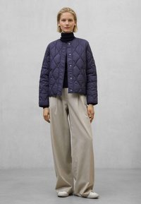 Marineblå quiltet jakke med rund krave og trykknapper, parret med lysebeige vide bukser og hvide sneakers. Glatte, bløde materialer.