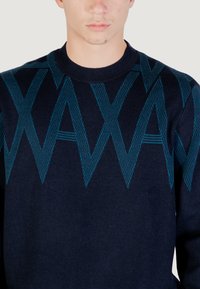 Homme portant un pull bleu marine avec un grand motif teal "WAX" tricoté sur la poitrine et les manches supérieures.