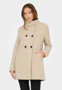 Manteau beige à double boutonnage avec des boutons noirs, col haut, poches latérales, tissu texturé et coupe décontractée.