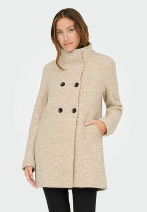 Manteau beige à double boutonnage avec des boutons noirs, col haut, poches latérales, tissu texturé et coupe décontractée.