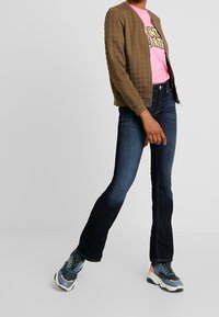 Bruine bomberjack met textuur, roze grafisch t-shirt, donkerblauwe uitlopende spijkerbroek en meerkleurige sneakers, die lagen van casual kleding laten zien.