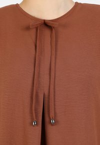 Bordeaux blouse met een ronde halslijn, voorzien van een strikdetail bij de hals en metallic accenten op de strikken. Zachte, gestructureerde stof.