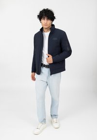 Schwarze Bomberjacke mit Stehkragen, Reißverschluss vorne, Seitentaschen und Ellbogenpatch-Details; kombiniert mit hellblauen Jeans und weißen Turnschuhen.