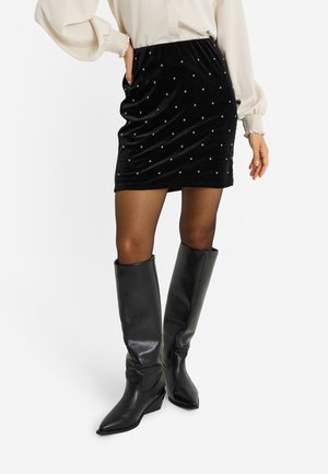 Mini falda de terciopelo negro con adornos metálicos en forma de diamante, combinada con botas negras de cuero hasta la rodilla con punta puntiaguda.