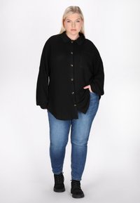 Camicetta nera con bottoni in tessuto testurizzato, con colletto e due tasche anteriori, abbinata a jeans blu e stivali neri stringati.