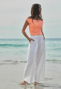 Top arancione fluo off-shoulder abbinato a pantaloni bianchi in lino a gamba larga, con tasche e un fit rilassato, in piedi su una spiaggia.