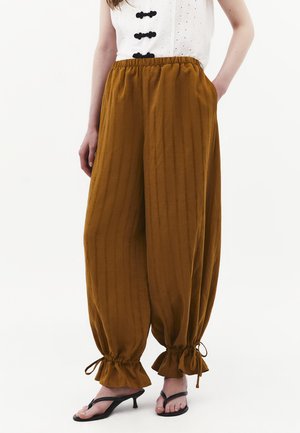 Bruna vida byxor med resår i midjan och volangprydda dragskoresår i muddarna, bärs med svarta sandaler med klack och vit topp med svarta knappar.