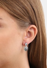 Sölvhängande örhängen med en rund stift som har en klar ädelsten, fästa vid en oval berlock med ett etsad mönster.