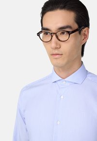 Šviesiai mėlynas dryžuotas marškinis su smailiu apykaklės kraštu, paprastais sagomis ir lengva medžiaga, derinamas su apvaliais žiniatinklio akinių rėmeliais.