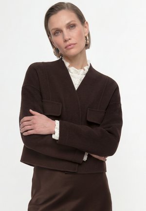 Mujer con el pelo corto que lleva una chaqueta marrón sobre una blusa blanca con volantes y pendientes de aro dorados, de pie con los brazos cruzados.