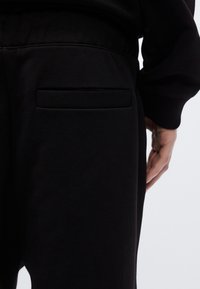 Μαύρο παντελόνι φόρμας με λεία υφή, που διαθέτει διακριτική πίσω τσέπη και ελαστική μέση. Κοντινή όψη από το πλάι.