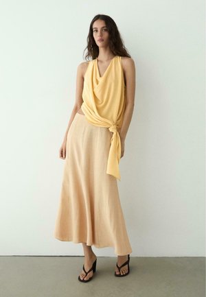 Vrouw draagt een gele mouwloze geplooide top gestrikt in de taille, beige midirok en zwarte sandalen met hak, staand tegen een effen witte muur.