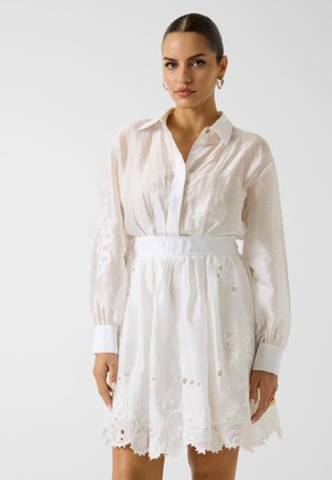 Femme aux cheveux plaqués en arrière portant une robe chemise blanche à manches longues avec des découpes florales brodées et des manches retroussées, debout devant un fond uni.
