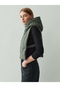 Grüne, gesteppte Weste mit Kapuze, die einen taillierten Schnitt und Seitentaschen aufweist, getragen über einem schwarzen Langarmshirt und dunklen Jeans.