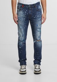 Blå denimjeans med slitna detaljer, splattermönster och revna knän. Har fem fickor och knappstängning. Bärs med svarta och vita sneakers.