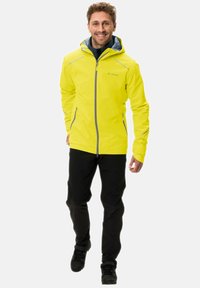 Helle gelbe wasserdichte Jacke mit grauen Akzenten, durchgehendem Reißverschluss, Kapuze und Reißverschlusstaschen, kombiniert mit schwarzen Hosen und robustem Schuhwerk.