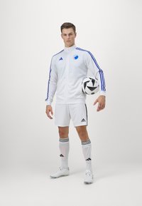 Bijeli Adidas trenirka s plavim prugama i timskim logotipom, u kombinaciji s kratkim hlačama, bijelim čarapama i kopačkama, drži crno-bijelu nogometnu loptu.
