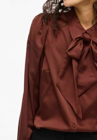 Brūns satīna blūze ar sasietu loku pie kakla, ar krokotām plecu detaļām un pogu aizdari apakšā. Gluda tekstūra, garas piedurknes.