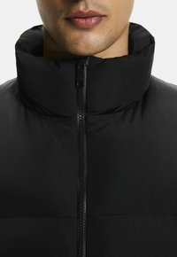 Chaqueta de plumas negra con cuello alto, que presenta un cierre frontal y una textura suave y mate. Sin patrones ni acentos visibles.