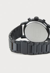 Montre en acier inoxydable noir avec un bracelet à maillons, un boîtier rond et des boutons-poussoirs texturés. Présente un dos lisse en argent avec gravure de logo.