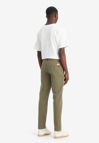Pantaloni verde oliva dal taglio slim, con due tasche posteriori e un piccolo etichetta del marchio. Abbinati a una camicia bianca a maniche corte.