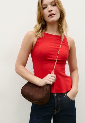 Junge Frau trägt ein ärmelloses rotes Oberteil und dunkle Jeans, hält eine kleine braune Umhängetasche mit einem goldenen Kettenriemen.