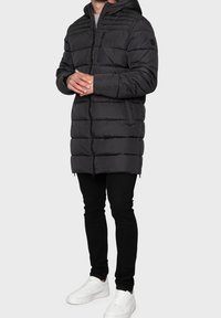 Doudoune noire à capuche, avec des sections matelassées, une fermeture éclair devant et une petite poche sur la poitrine. Associée à un pantalon noir et des baskets blanches.