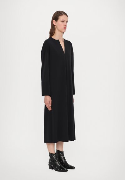 Crna haljina dugih rukava s dubokim V-izrezom i slobodnim krojem, izrađena od glatkog materijala. Uparena s crnim čizmama do gležnja s dodatkom rupica.