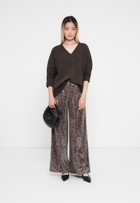 Bruine oversized trui gecombineerd met wijde, met pailletten bedekte broek in grijs- en bronsnuancen, aangevuld met een zwarte handtas en puntige schoenen.