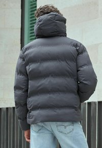 Tamsiai pilkas pūkinis švarkas su aukšta apykakle ir siuvinėtu dizainu. Suderintas su šviesiai mėlynais džinsais, prieš neutralų foną.