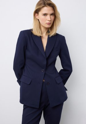 Blonde vrouw in een op maat gemaakt marineblauw pak met handen achter haar rug, staand tegen een effen lichtgrijze achtergrond.