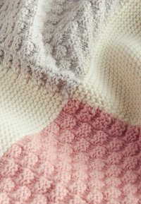 Knitted throw blanket featuring a textured pattern. Colors: light gray, cream, and soft pink. Composed of thick, cozy yarn.
