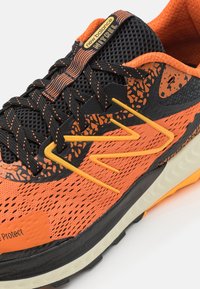 Zapatilla de running naranja y negra con parte superior de malla, acentos texturizados y logo amarillo. Presenta un patrón elevado de manera prominente en el costado.