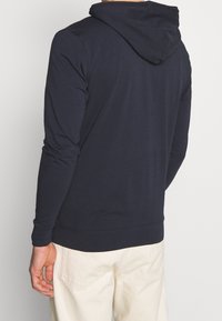 Tmavě modrá mikina s dlouhým rukávem, kapucí se stahovací šňůrkou, žebrovanými manžetami a hladkým povrchem, nošená s světlými kalhotami.