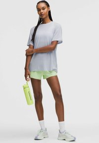 Vrouw met ingevlochten haar, draagend een lichtgrijze T-shirt, limoen groene shorts, witte sokken, sneakers en een limoen groene Lululemon waterfles.