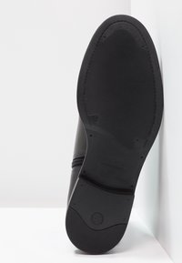 Zapato de cuero negro con una superficie suave, suela de goma texturizada y un detalle de cremallera. Cuenta con la marca de la empresa y una etiqueta de tamaño.
