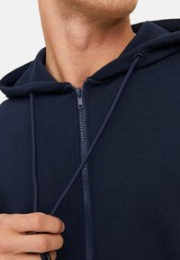 Marineblauwe zip-up hoodie met een trekkoordcapuchon, gemaakt van zacht materiaal, voorzien van een gladde rits en een gestructureerde afwerking.